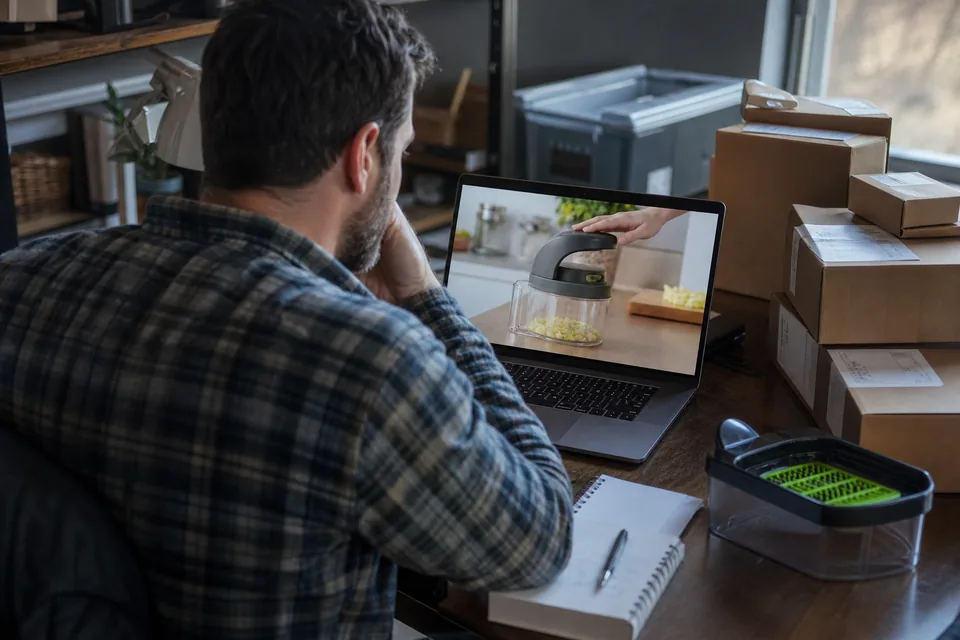 Amazon seller at a home office desk watching a creator's product demo video on a laptop next to a stack of shipping boxes
