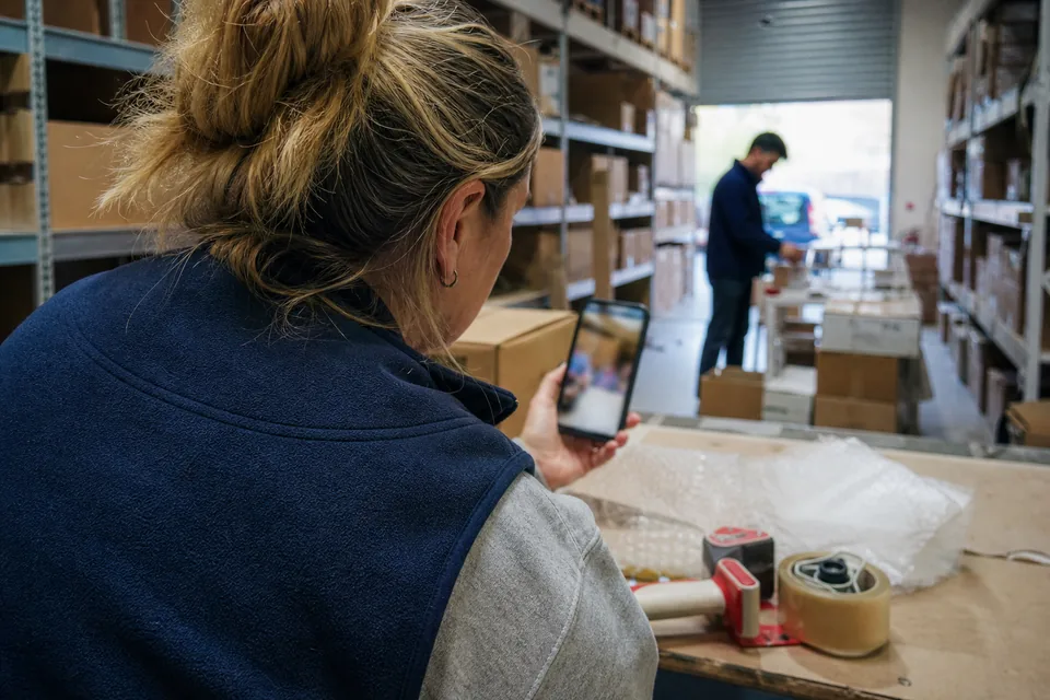 Ecommerce manager in a small warehouse watching a creator's vertical video on her phone next to a packing table