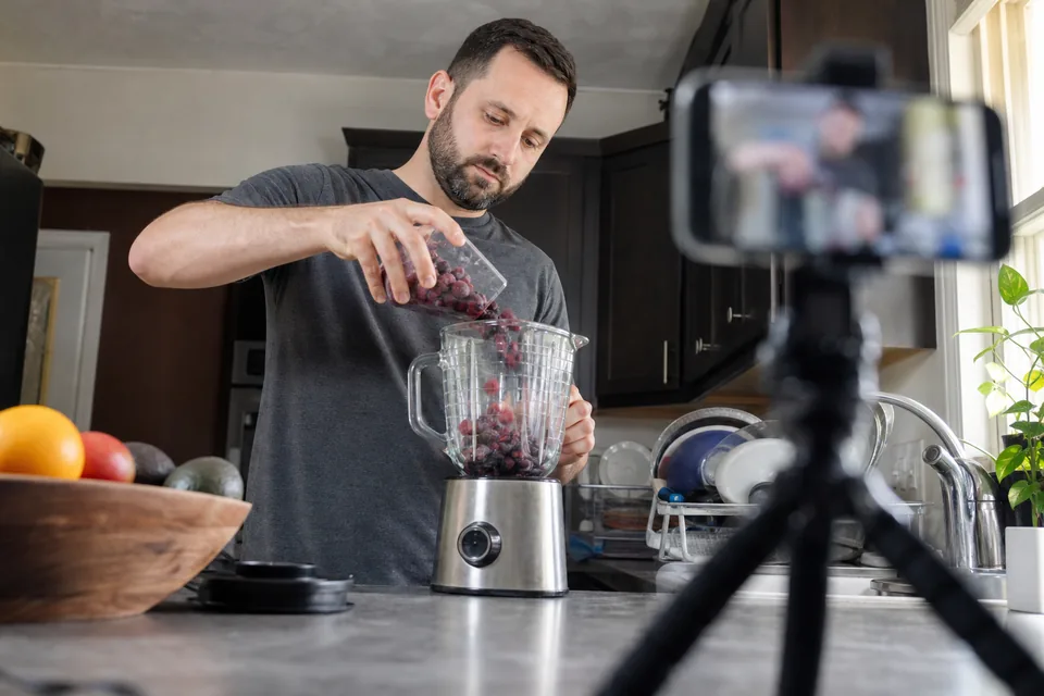 Adult creator demonstrating a blender in a home kitchen while a phone on a tripod films from counter height