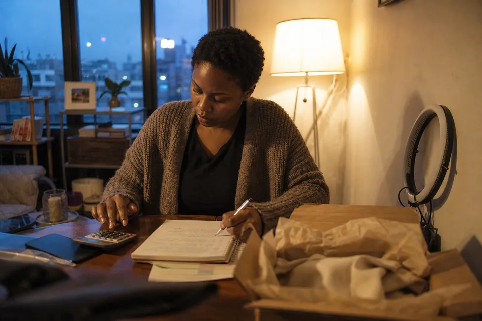 Adult creator at her kitchen table working out a quote with a calculator and a notebook, a shipped product box beside her
