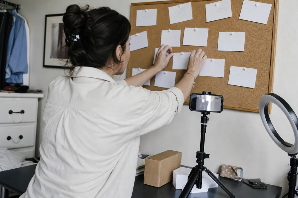 Adult creator in a home studio pinning index cards with shoot plans to a corkboard above her desk