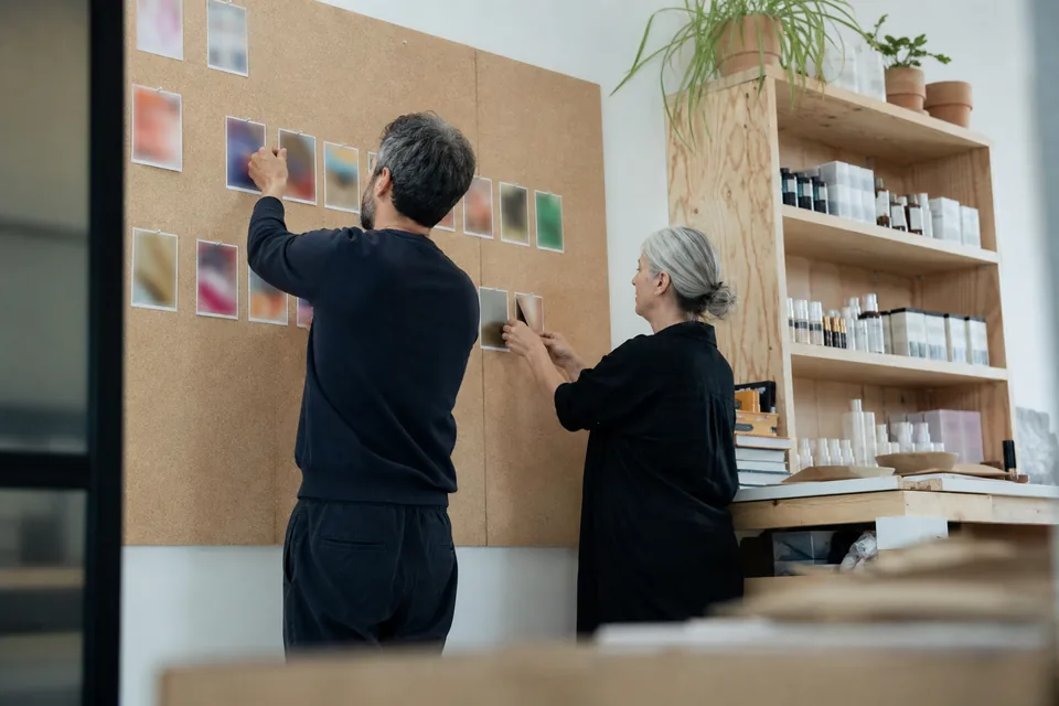Two marketers at a small brand pinning printed video stills onto a corkboard wall to plan next month's ads