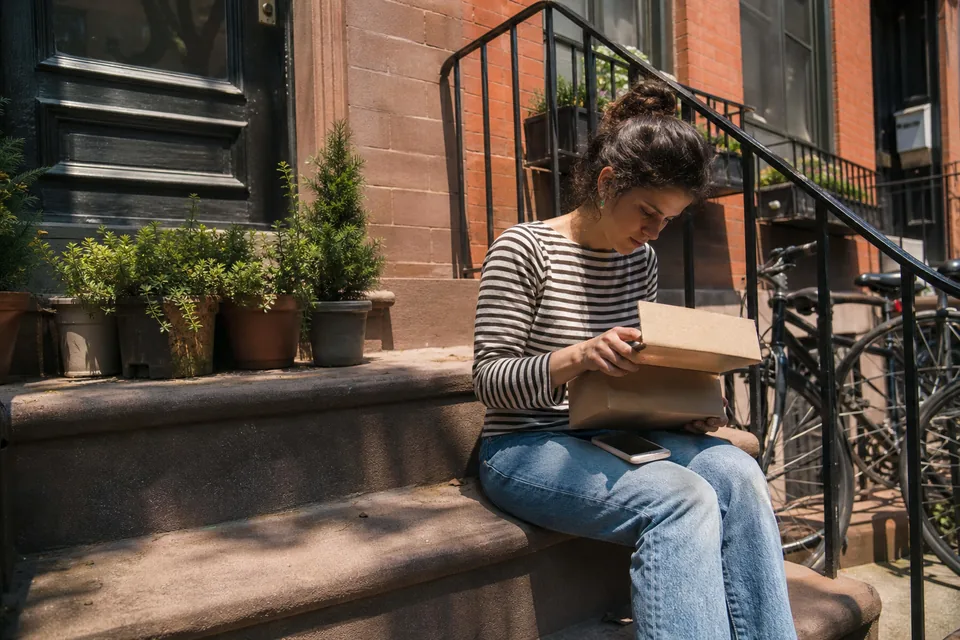 Adult creator opening a shipped product parcel on the steps outside her apartment building, phone in her other hand