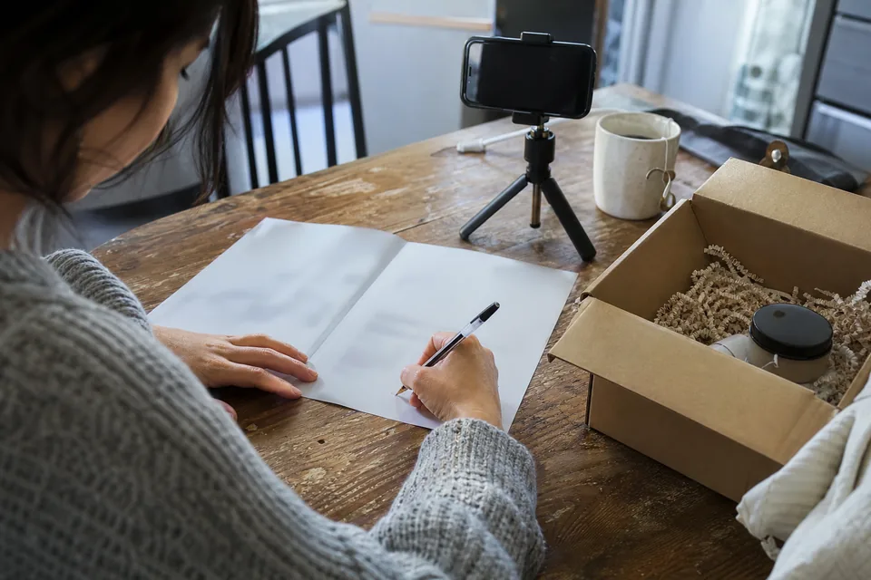 Creator's hand signing a printed campaign agreement with a ballpoint pen at a kitchen table, the shipped product box open beside it and a phone on a small tripod ready to film