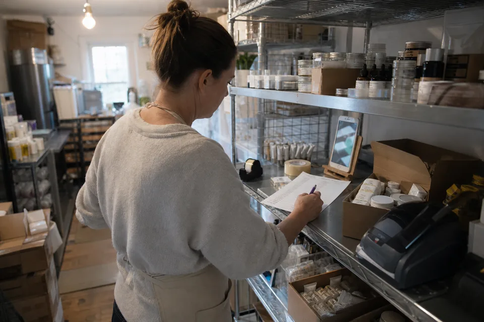 Owner of a small skincare brand in her shop's back room ticking off delivered creator videos on a printed checklist, a phone playing a vertical video on a stand beside cartons of samples